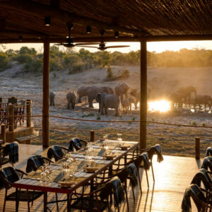 Tables laid on the open dining deck at Savute Safari Lodge with elephants at the waterhole beyond, Chobe National Park, Botswana