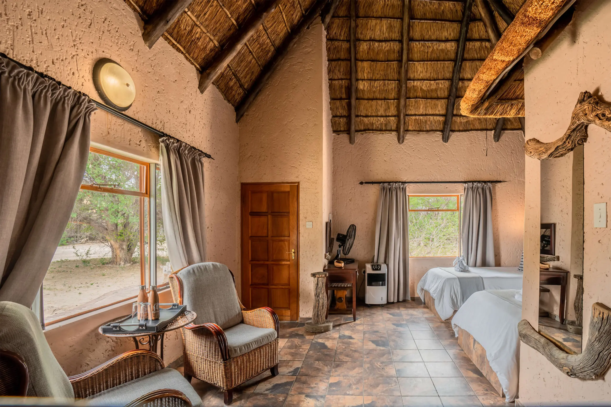 A chalet bedroom under a high thatched roof with windows onto the Kalahari bush at Grassland Safari Lodge, Botswana