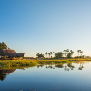 The tented rooms and thatched main area of Moremi Crossing reflected in a Delta channel at sunset, Okavango Delta, Botswana